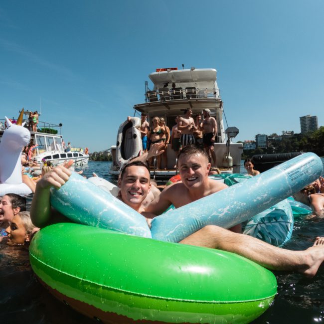 A group of people enjoying a sunny day on the water with inflatable rafts and boats. Two individuals in the foreground smile while holding onto a large green and blue inflatable, creating the perfect scene for a Sydney boat party hire.