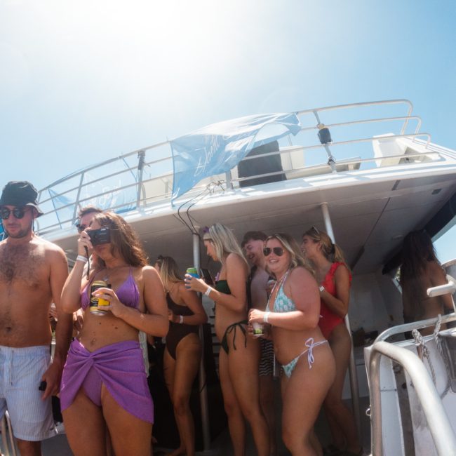 A group of people in swimwear are standing and sitting on a boat under the sun, some holding drinks, enjoying a lively Sydney boat party hire.