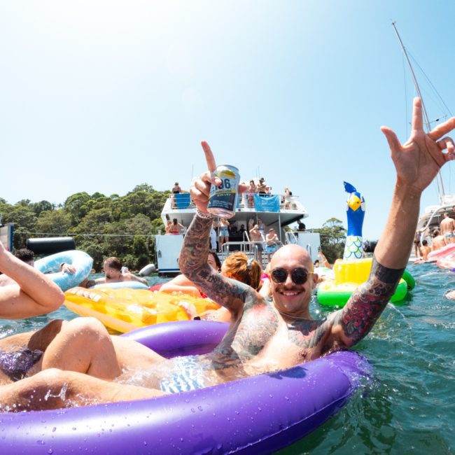 People floating on inflatables and swimming in a body of water, with boats and more inflatables in the background. One person in a purple inflatable is holding a can and gesturing with their hand, enjoying the Catamaran party Sydney vibes.