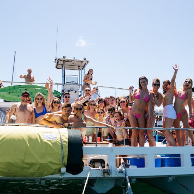 A group of people enjoying a sunny day on a private yacht charter in Sydney Harbour, wearing swimwear and sunglasses. Some are standing, while others are sitting or posing for the photo.