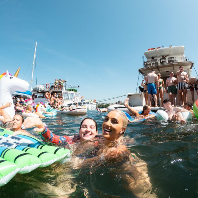 A group of people enjoy a sunny day in the water, surrounded by inflatable floats and boats, with some on the boats and others swimming or floating. The lively scene features a Catamaran party Sydney, adding to the festive atmosphere.