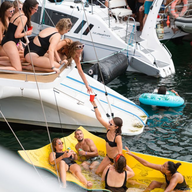 A group of people in swimsuits socialize on a private yacht charter in Sydney Harbour and a floating mat in the water, with some reaching out to each other. Other boats and inflatables are visible in the background, making for an unforgettable catamaran party Sydney experience.