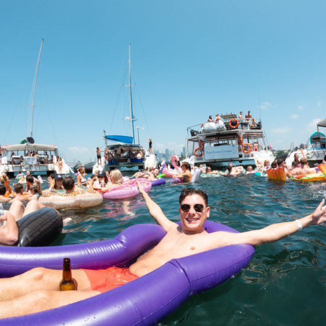 A person in sunglasses lounges on a purple float in the water, arms outstretched, with a beer bottle nearby. Several boats and other people on floats are enjoying a sunny day, including a private yacht charter on Sydney Harbour adding to the vibrant atmosphere.