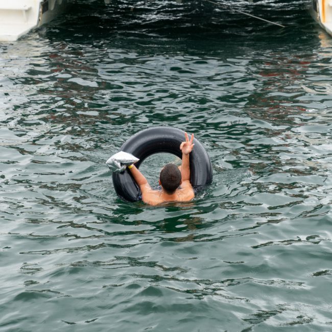 A person is afloat in water using a tire as a flotation device, holding a drink in one hand and making a peace sign with the other. Nearby, the excitement of a Sydney boat party hire adds to the vibrant summer day.