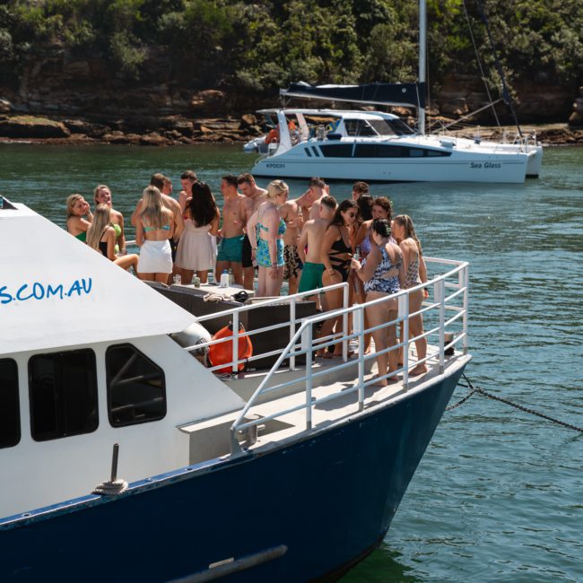 A group of people is standing on the deck of a luxury yacht hire Sydney in the water. In the background, another boat and a wooded shoreline are visible.