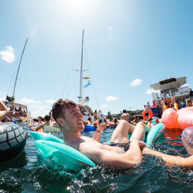 People are floating on inflatable rafts in the water, with boats and clear skies in the background. A man in the center is holding a drink and smiling. A large flamingo inflatable is also visible, reflecting the fun of a Sydney boat party hire.