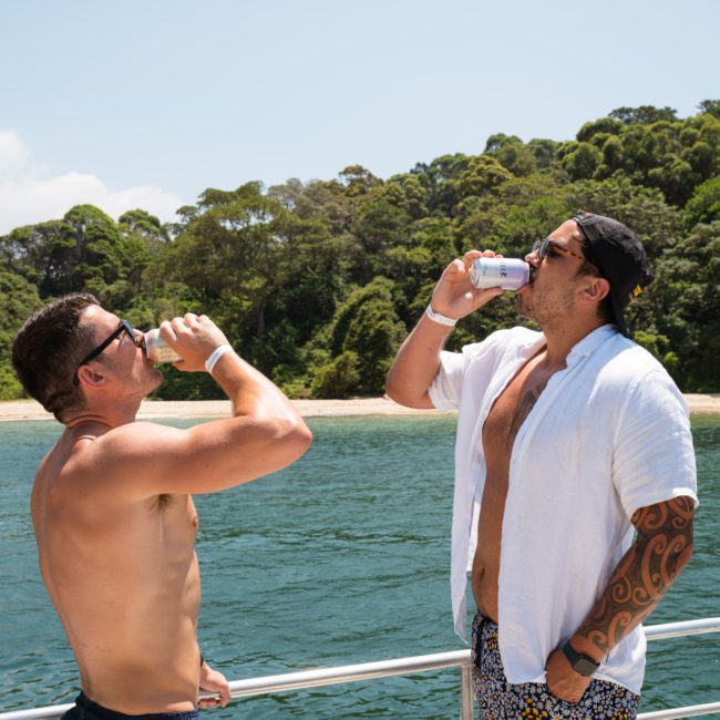 Two men standing on a boat deck, drinking canned beverages, with a scenic view of trees and water in the background, enjoying their luxury yacht hire in Sydney.