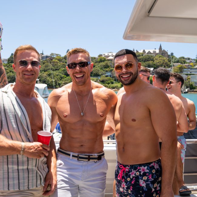 Three men standing on a boat deck on a sunny day, smiling at the camera during a Sydney boat party hire. The man in the center is shirtless, while the other two wear shirts. Several people and a shoreline can be seen in the background.