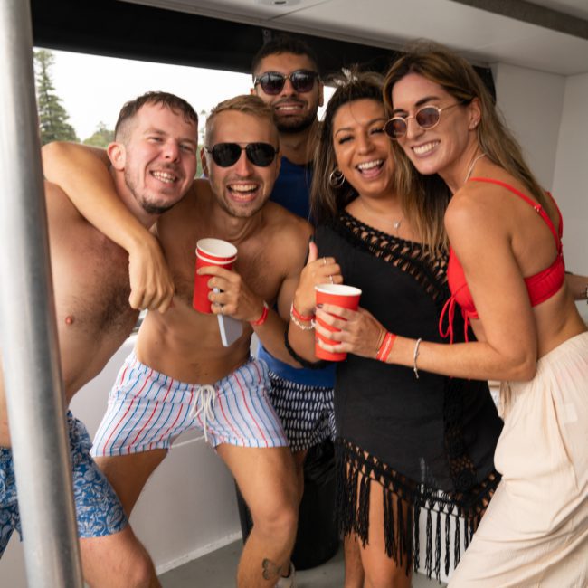 A group of five people smiling and posing for a photo on a boat in Sydney Harbour. They are dressed in casual, summer attire, and two of them are holding red cups, enjoying what seems to be a fantastic Sydney boat party hire.