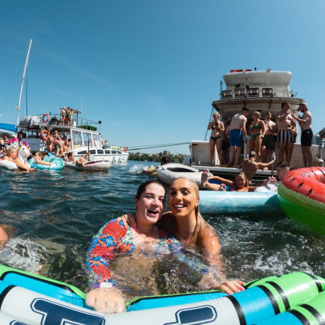People floating on inflatable rafts and swimming near large boats during a sunny water party on a private yacht charter in Sydney Harbour.
