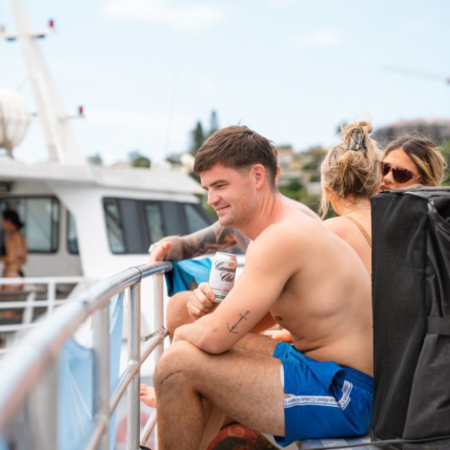 A shirtless man in blue shorts sits on the deck of a boat, holding a drink can. Several other people are also on the boat, enjoying a sunny day. It looks like they're part of a private yacht charter Sydney Harbour experience.