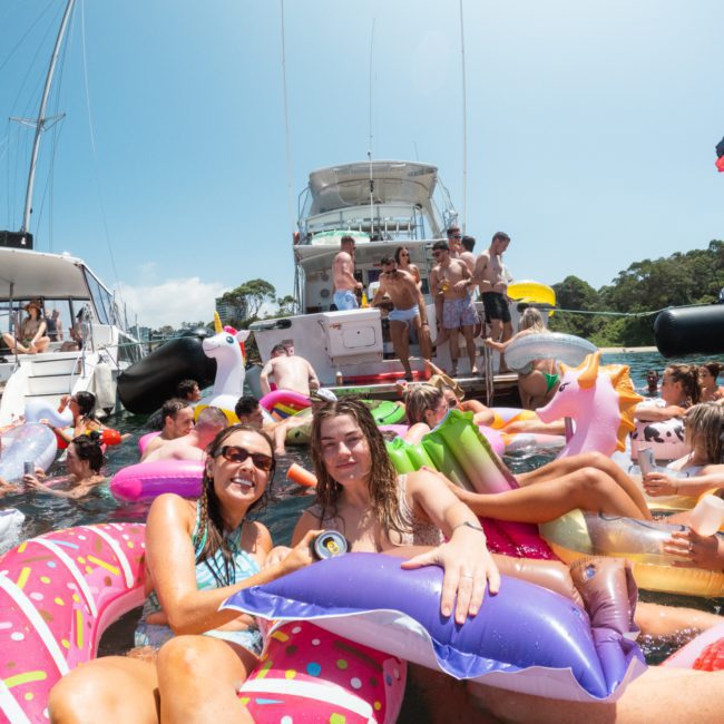 A group of people enjoy a sunny day on a variety of inflatable pool floats in the water near two luxurious yachts, perfect for any Sydney boat party hire or DJ boat hire Sydney.