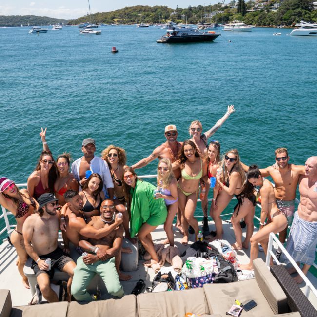A group of people are gathered on the deck of a boat in swimwear, posing and smiling for a photo with a scenic water and boat-filled background, perfect for a Sydney boat party hire.