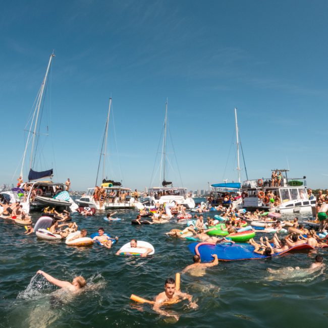 A bustling scene of people swimming and lounging on inflatables in the water, surrounded by boats under a clear blue sky—an ideal setting for a Sydney boat party hire.