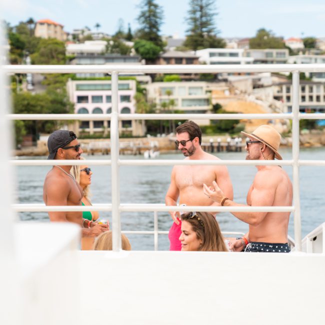 A group of people in swimwear are having a conversation on a luxury yacht hire in Sydney, with coastal residential buildings visible in the background.