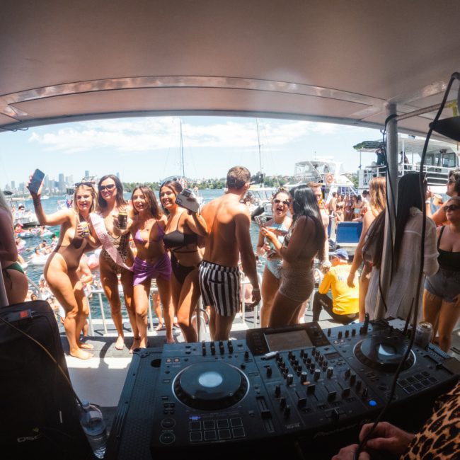 People gathered on a boat deck, enjoying a sunny day with drinks. A DJ is playing music, and a group in swimsuits is posing for a photo. Water and boats are visible in the background during this private yacht charter Sydney Harbour event.