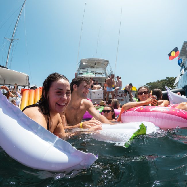 Group of people in water with inflatable toys near a boat on a sunny day, enjoying a DJ boat hire Sydney experience.