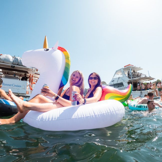 Two people sit on a large inflatable unicorn float in a body of water, holding cans. Other people and boats are visible in the background under a clear sky, with the elegance of luxury yacht hire Sydney enhancing the scene.