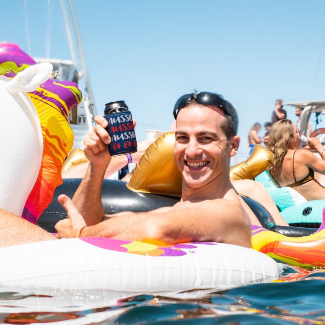 A man wearing sunglasses is sitting on an inflatable unicorn in the water, holding a beverage can cover. Other people and luxury yacht hires Sydney are visible in the background.