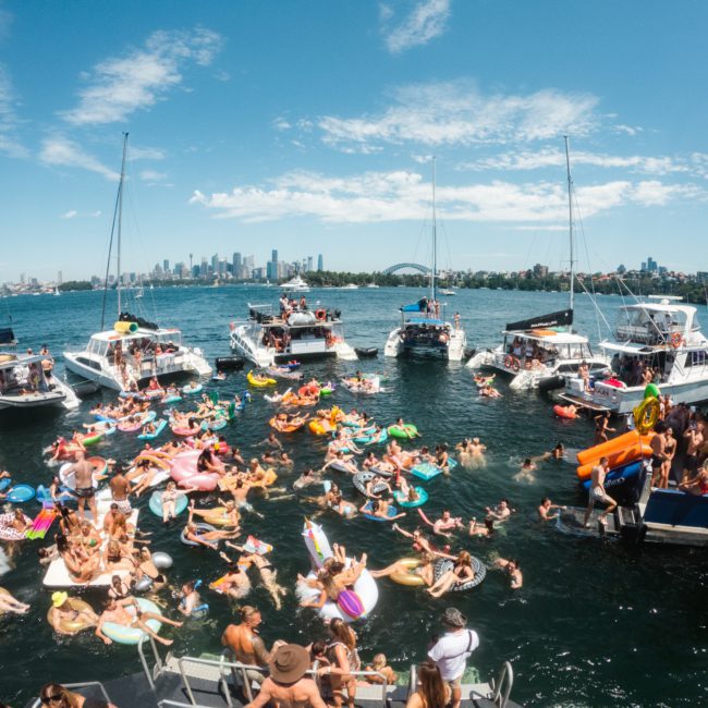 Dozens of people on inflatables gather in the water near anchored boats on a sunny day with the city skyline in the background, enjoying a luxurious atmosphere. Perfect for those considering Luxury yacht hire Sydney or Corporate boat events Sydney.