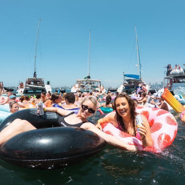 People are enjoying a sunny day on the water, floating on inflatable tubes with boats anchored in the background. The two women in the foreground are smiling and holding drinks, making it the perfect scene for a Catamaran party Sydney or Sydney boat party hire event.