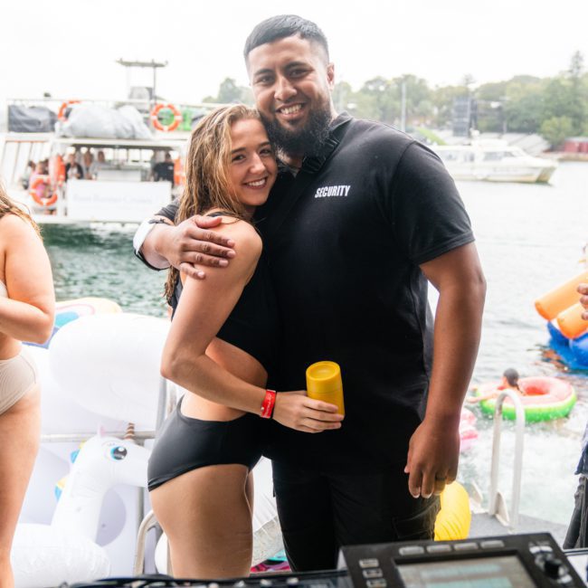 A woman in a black bikini and a man in a black "Security" shirt hug on a boat during an energetic Sydney boat party hire. The background shows people and inflatables on the water, while DJ equipment is visible in the foreground.