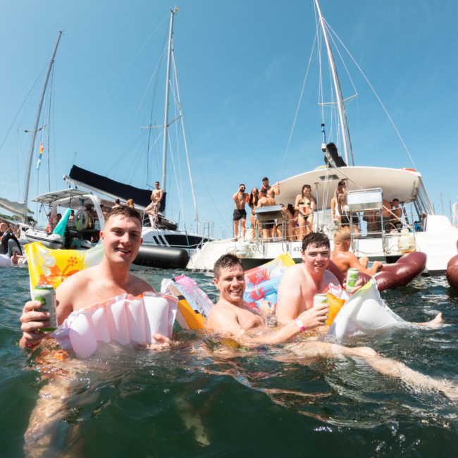 Three people in a lake hold inflatable toys and drinks, with several others and boats in the background on a sunny day, enjoying a luxury yacht hire Sydney experience.