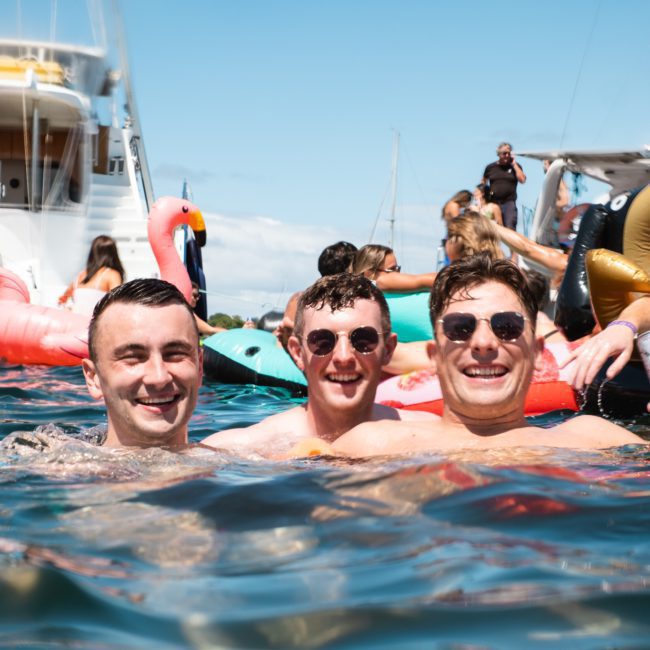 Three men are smiling and standing in the water, surrounded by floaties and other people with boats in the background under a sunny sky, enjoying what looks like a fantastic Sydney boat party hire.