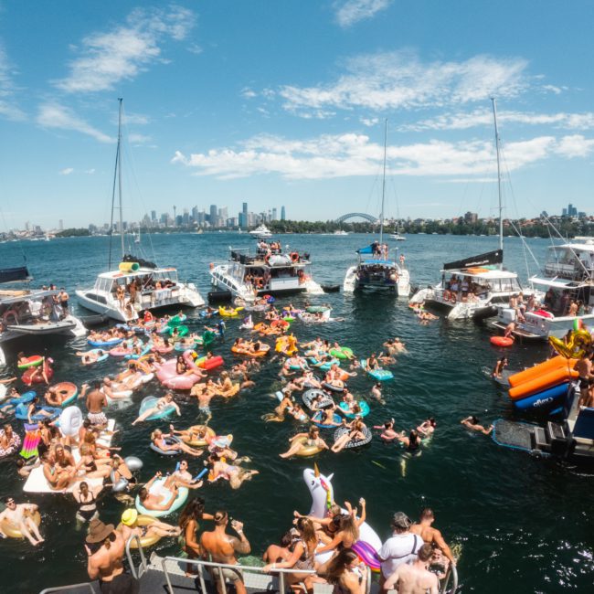 A lively aquatic gathering featuring numerous people on inflatable rafts and boats, enjoying a sunny day on the water with a cityscape in the background, alongside luxury yacht hire in Sydney.