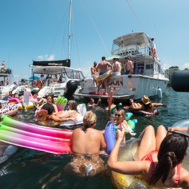 A group of people relax and socialize in inflatable floats and on boats during a sunny, crowded Sydney boat party hire.