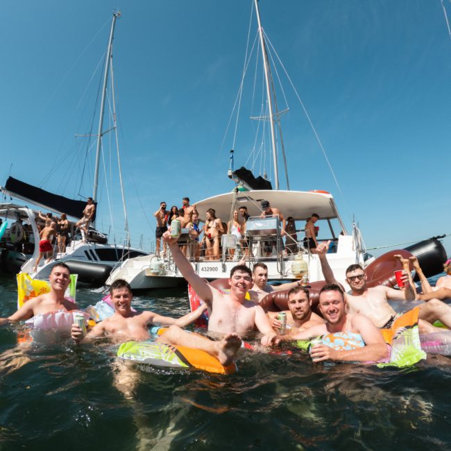 A group of people are enjoying a sunny day on the water, floating on inflatables near boats. They are smiling and holding drinks, embodying the perfect atmosphere for a Sydney boat party hire. More people are visible on the boats in the background.