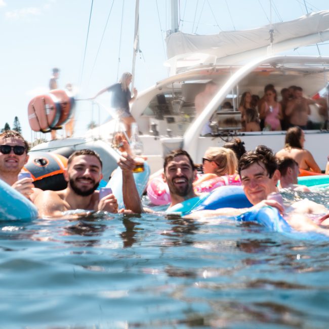 Four people smiling and posing in the water with floating devices near a boat in a sunny, outdoor setting. More people can be seen enjoying themselves on the boat, perfect for corporate boat events Sydney.