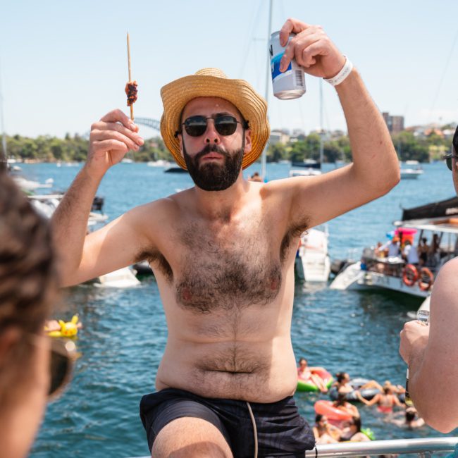 A man wearing a straw hat and sunglasses holds a drink can and a skewer on a luxury yacht hire in Sydney. Other boats and people are visible in the background on a sunny day.