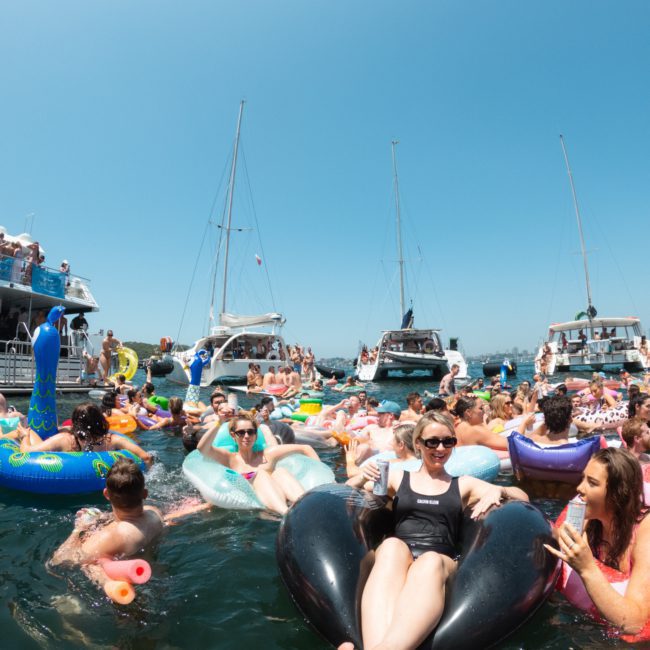 People float on inflatable rafts and swim near boats in a crowded, sunny, waterfront gathering. Celebrate on the water with a private yacht charter Sydney Harbour or enjoy music with DJ boat hire Sydney for an unforgettable experience.