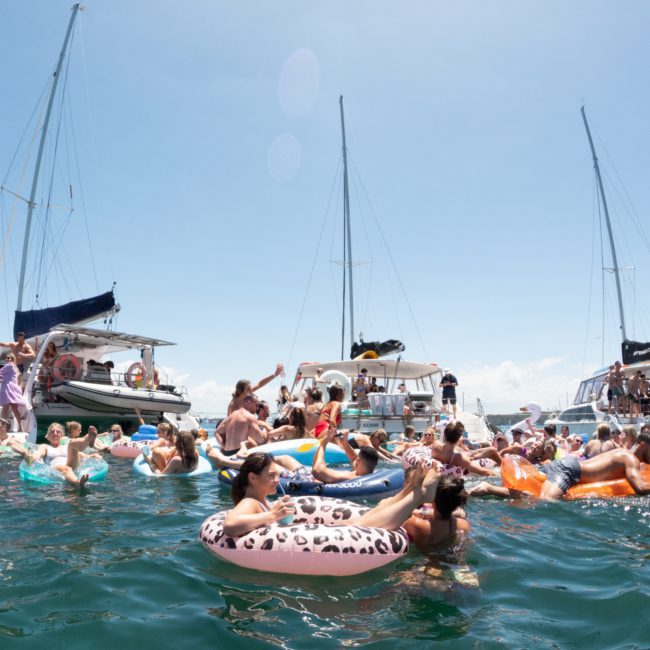 A group of people floats on inflatable tubes in the water between several anchored boats on a sunny day, enjoying a Private yacht charter Sydney Harbour.