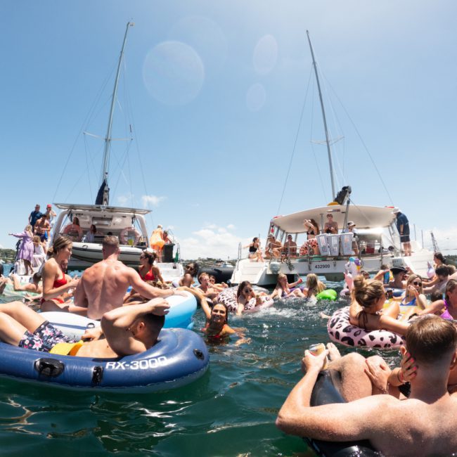 A group of people on various inflatables in the water, surrounded by several boats during a sunny day, enjoying a private yacht charter on Sydney Harbour.