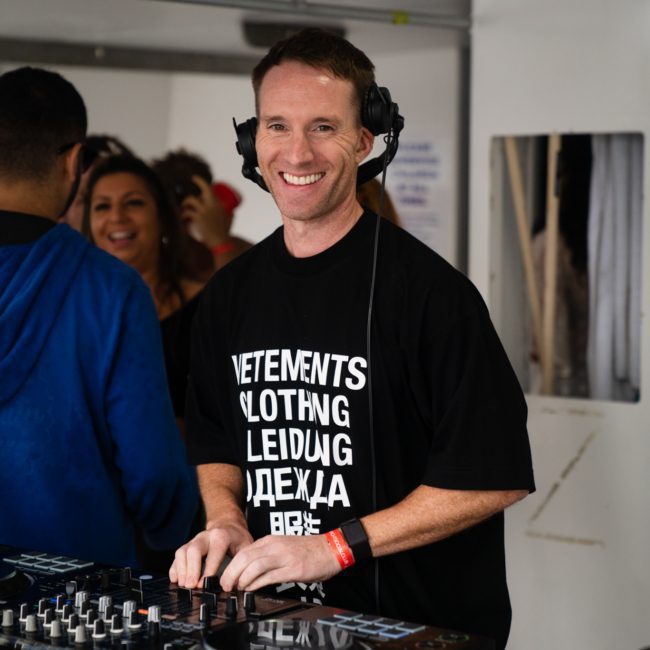 A DJ wearing a black T-shirt and headphones stands behind a mixing console, smiling at the camera. People in the background are engaged in conversation during a lively Sydney boat party hire.