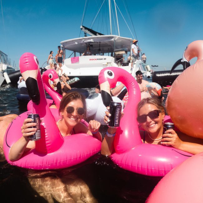 Two women on pink flamingo pool floats hold canned drinks while smiling at a catamaran party in Sydney with several other people and boats in the background.