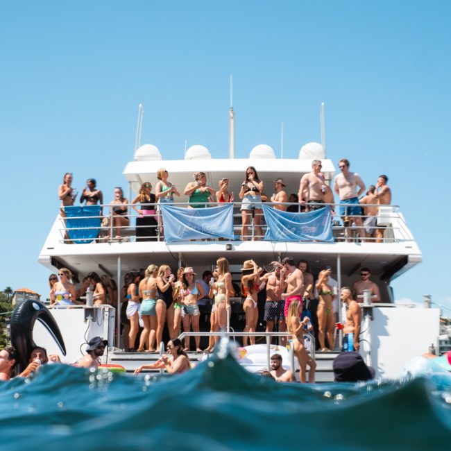 A large group of people is gathered on a boat, with some swimming in the water nearby. The boat is docked near a shoreline under a clear blue sky, perfect for a Sydney boat party hire.