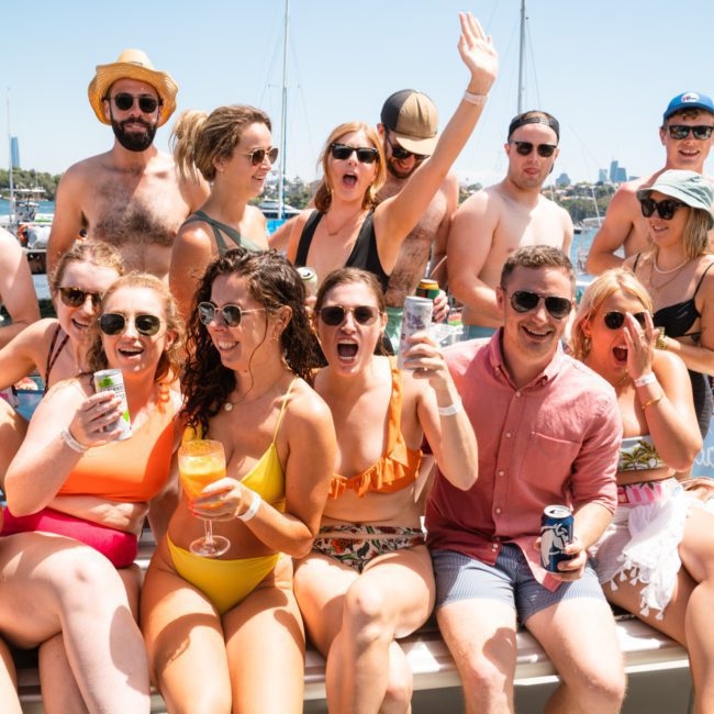 A group of people in swimwear enjoying a sunny day on a luxury yacht hire in Sydney, with a city skyline in the background. Some individuals are holding drinks and appear to be laughing and smiling.