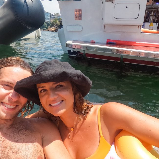 A man and woman are floating on an inflatable device in the water near a boat. The woman is wearing a black hat and a yellow bikini. In the background, another person is seen sitting on the boat, enjoying their time during a Catamaran party in Sydney.