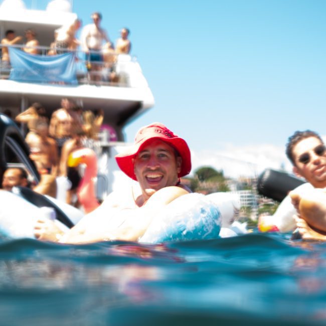 People floating on inflatable tubes in the water, with a crowded boat in the background. One person in a red hat is prominently smiling at the camera. It's an ideal scene for a Sydney boat party hire, where fun and relaxation go hand in hand.