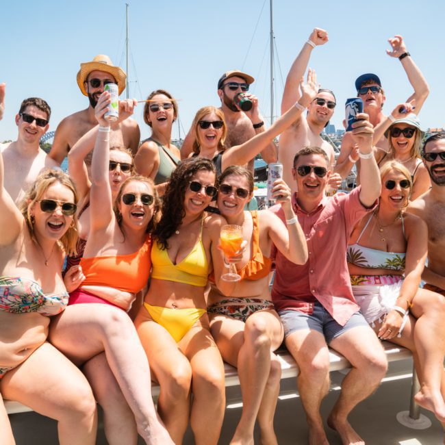 A group of people in swimwear are gathered on a luxury yacht in Sydney, smiling and raising their drinks, with a marina and other boats in the background.