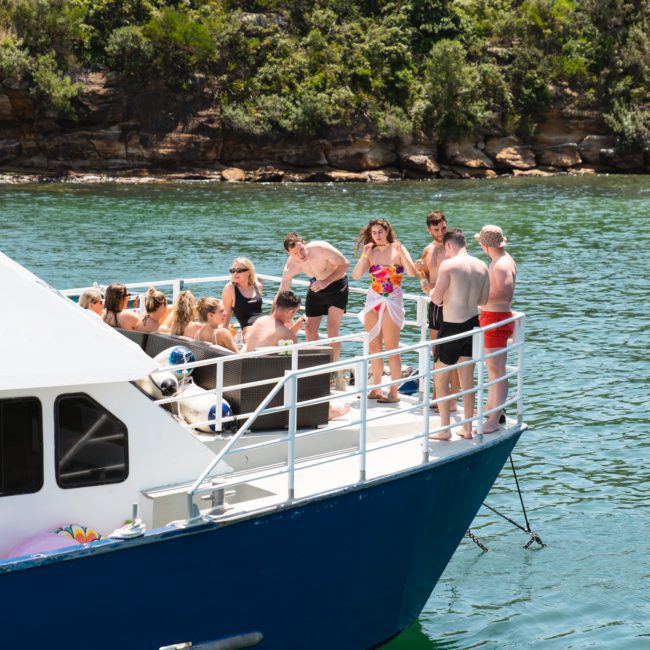 A group of people are gathered on the deck of a catamaran party Sydney, cruising on a body of water surrounded by a rocky, tree-covered shoreline.