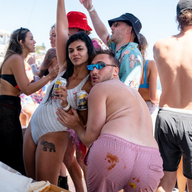A group of people is enjoying a party outdoors, with some holding drinks. One person in a white outfit and another in pink shorts are posing together. Other individuals are socializing in the background, reminiscent of a lively Sydney boat party hire or a fun-filled catamaran party Sydney event.