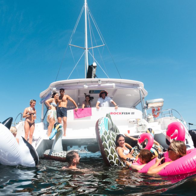 A group of people enjoy a sunny day on and around a boat named "Rockfish III," surrounded by inflatable pool floats in various shapes, including swans and an orca. Perfect for those seeking a lively Sydney boat party hire experience.