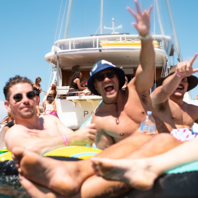 A lively group of people on inflatable pool floats are having a fantastic time in the water, with a luxury yacht hire Sydney in the background. Some are posing and cheering under the sunny sky.