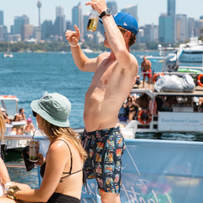A shirtless man holding a drink raises his arms while standing on a boat. Nearby, a woman in a hat and bikini sits relaxing. The city skyline and other boats are visible in the background, capturing the essence of a Sydney boat party hire or luxury yacht hire Sydney experience.