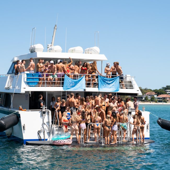 A large group of people in swimwear are gathered on a two-level yacht, some on the deck and others on the swim platform, enjoying a sunny day near the shore with a DJ boat hire Sydney.