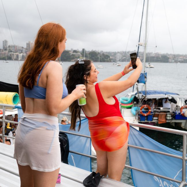 Two women in swimsuits are taking a selfie on a boat deck with a marina and other boats in the background. One of them is holding a drink can. The day appears overcast, perfect for enjoying a luxury yacht hire in Sydney.